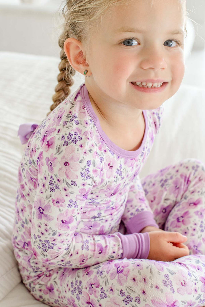 A smiling girl with a braid glancing over her shoulder, wearing a Caden Lane cream knit pajama set in a Lilac Dreams print featuring watercolor lilac flowers.