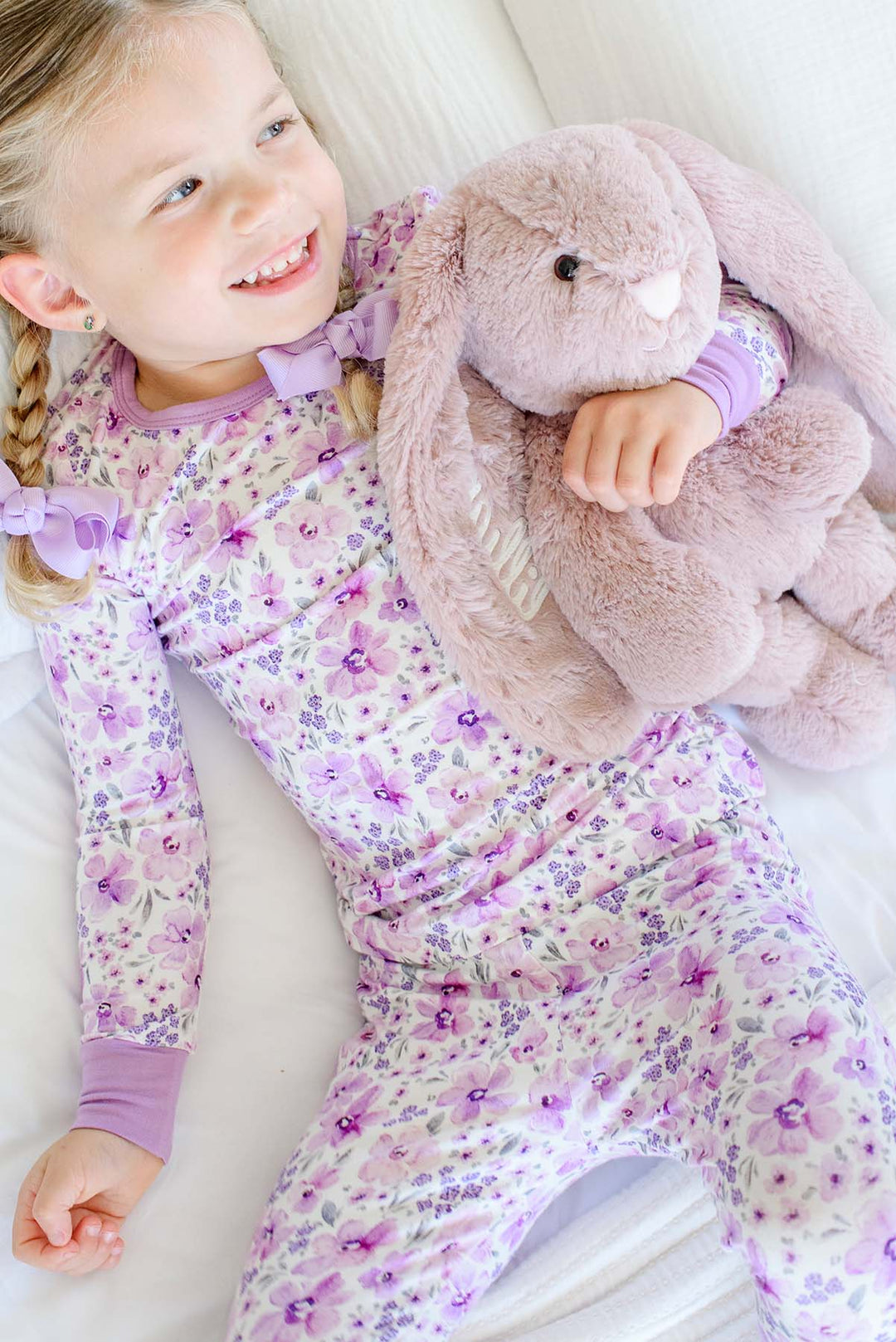A laughing girl lying on white bedding hugging a plush bunny, wearing a Caden Lane cream knit pajama set in a Lilac Dreams print featuring watercolor flowers.