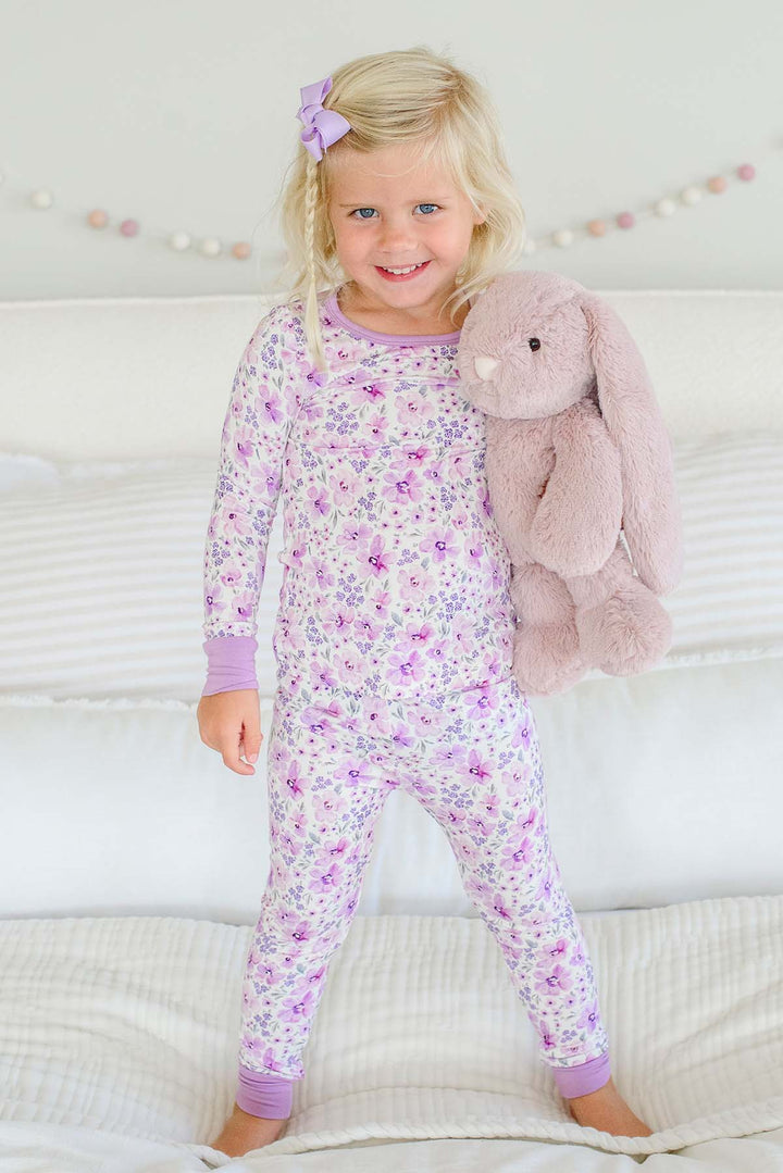 A blonde girl standing on a bed wearing a Caden Lane cream knit pajama set in a Lilac Dreams floral print, paired with a lilac hair bow and plush bunny.