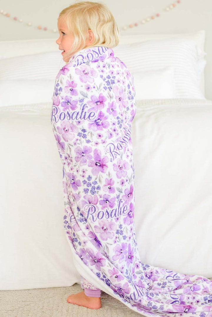Back view of a smiling blonde toddler standing by a bed, wrapped in a Caden Lane white plush blanket in a Lilac Dreams print featuring purple watercolor flowers, reading "Rosalie."