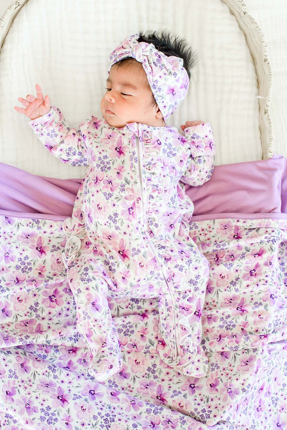 Overhead shot of a newborn in a wicker bassinet wearing a Caden Lane white knit ruffle footie in a Lilac Dreams print featuring purple flowers, paired with a matching blanket.