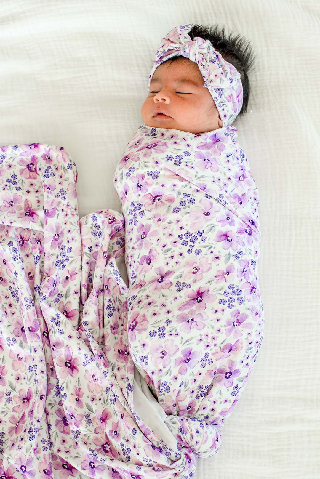 Full-length view of a sleeping newborn wearing a Caden Lane white knit swaddle in a Lilac Dreams print featuring purple flowers and grey leaves, paired with a matching headwrap.