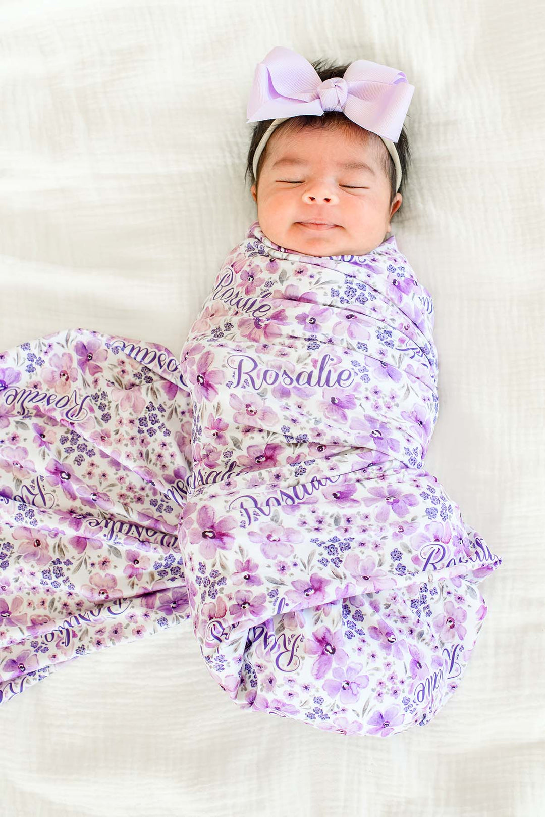 Overhead shot of a smiling, sleeping newborn on white bedding wearing a Caden Lane white knit swaddle in a Lilac Dreams print featuring lavender blooms and grey leaves, reading "Rosalie."