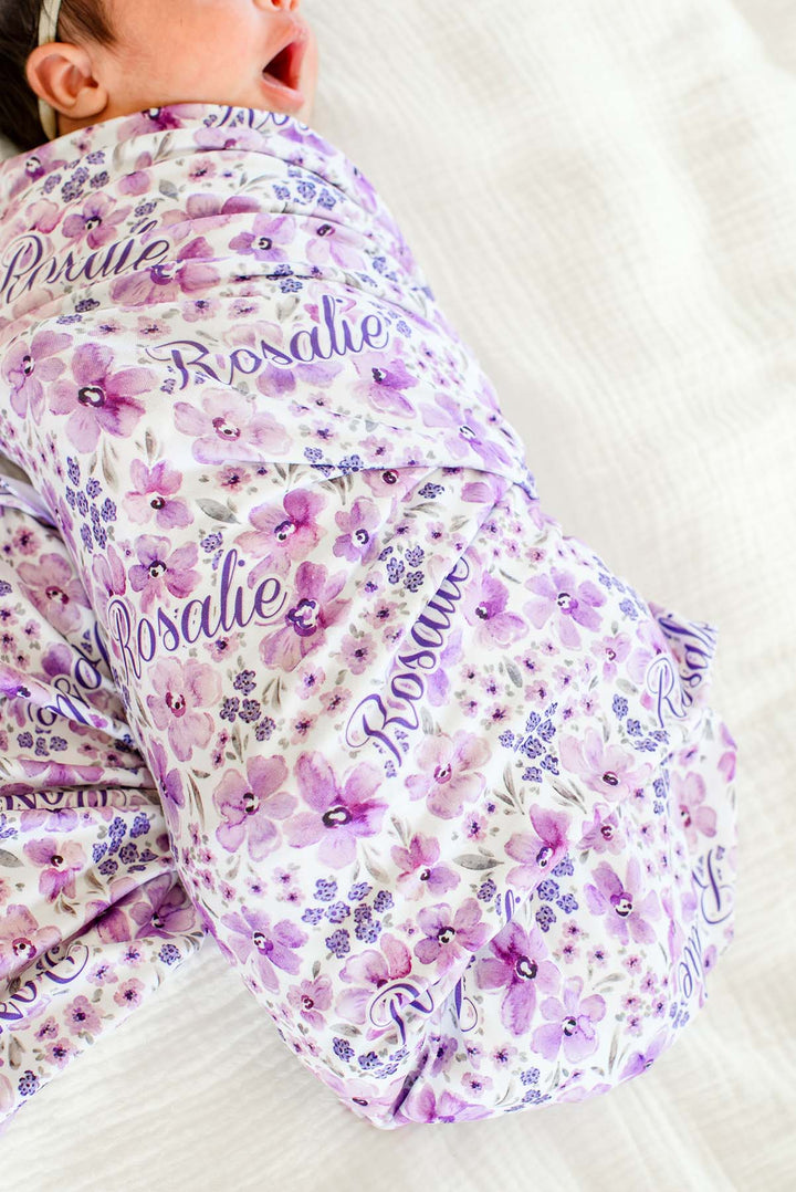 Close-up of a yawning newborn, face cropped, wearing a Caden Lane white knit swaddle in a Lilac Dreams print featuring pink and purple watercolor blooms, reading "Rosalie."