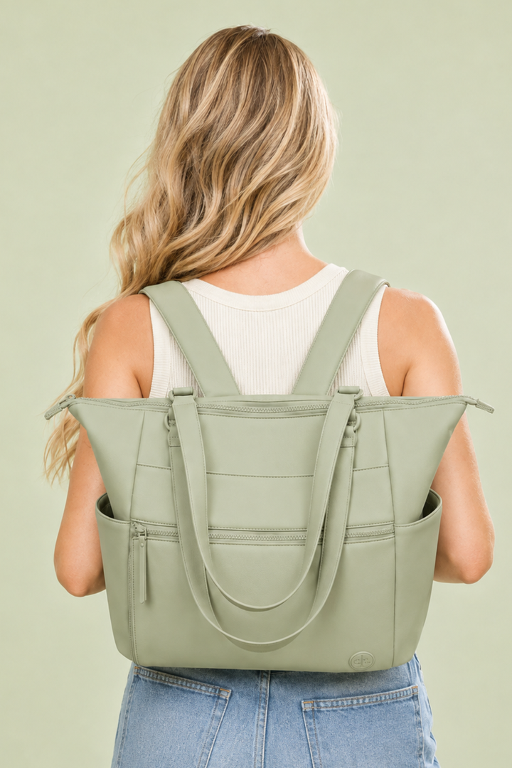 A woman viewed from behind wearing a sage green backpack-style tote bag over her shoulders, paired with a white ribbed tank top and light blue jeans, against a solid sage green background.