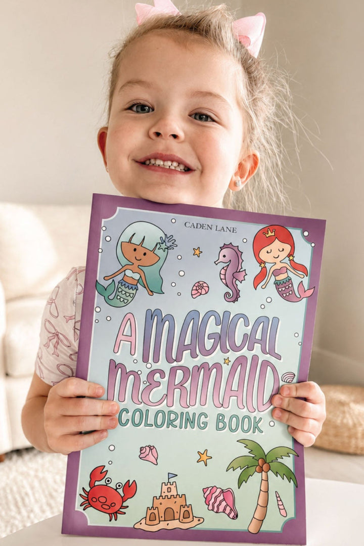 Smiling child wearing mermaid-fin hair clips holding the Caden Lane "A Magical Mermaid" coloring book.