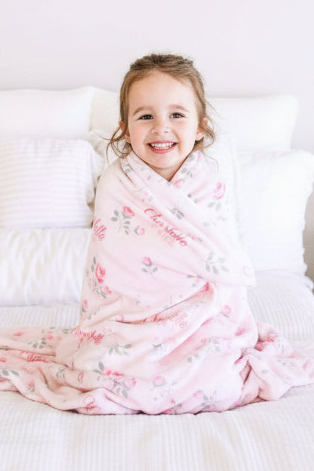 Caden Lane personalized baby blanket, toddler sitting on bed wrapped in Rose Garden pink floral blanket with custom name