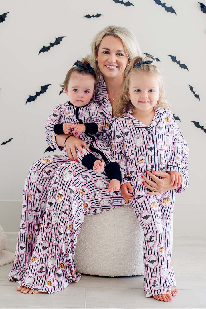 A woman and two young girls wearing matching Halloween-themed pajamas with a purple, white, and orange witch pattern, sitting together on a white round ottoman against a white wall decorated with black bat silhouettes.