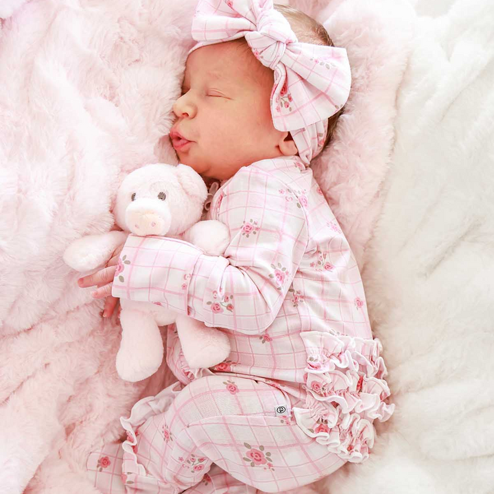 Sleeping newborn hugging a plush pig, wearing a Caden Lane pink bamboo ruffle zipper footie in a Primrose Plaid print featuring plaid and roses, paired with a matching bow headwrap.