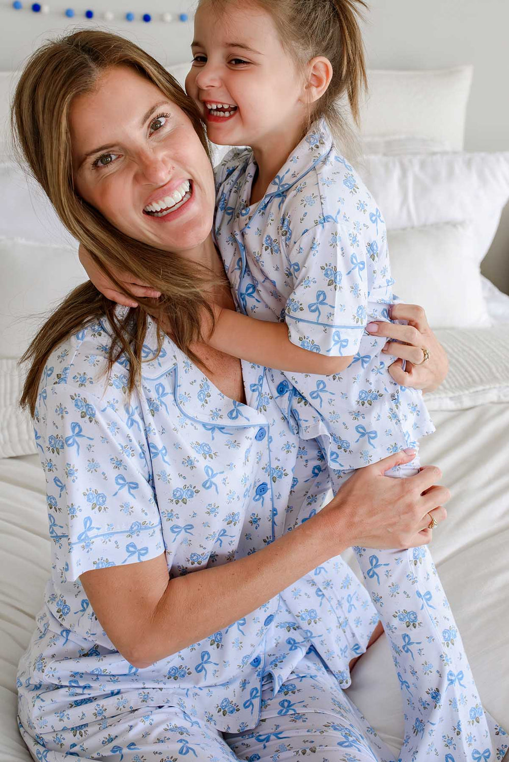 Mother and daughter smiling and hugging, both wearing matching Tied In Blooms bamboo lounge sets in blue floral and bow print