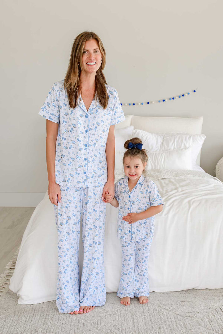 Mother and daughter standing side by side holding hands by a bed, both wearing matching Tied In Blooms bamboo lounge sets in blue floral and bow print