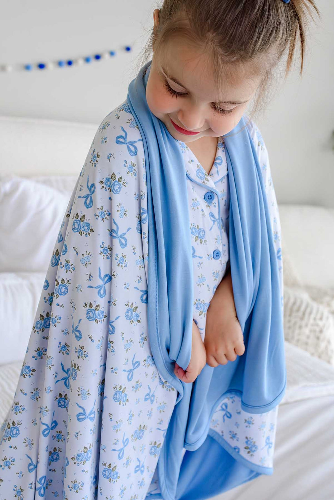Girl wrapped in a Tied In Blooms double sided bamboo blanket with blue floral and bow print, looking down