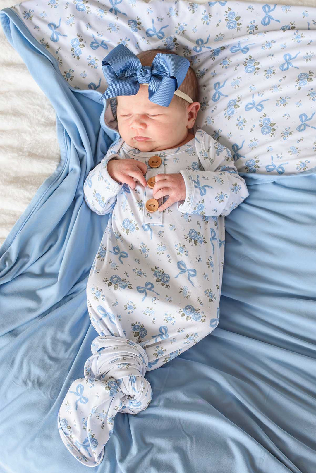 Sleeping newborn baby in a Tied In Blooms knot gown with blue floral and bow print, wearing a blue bow headband