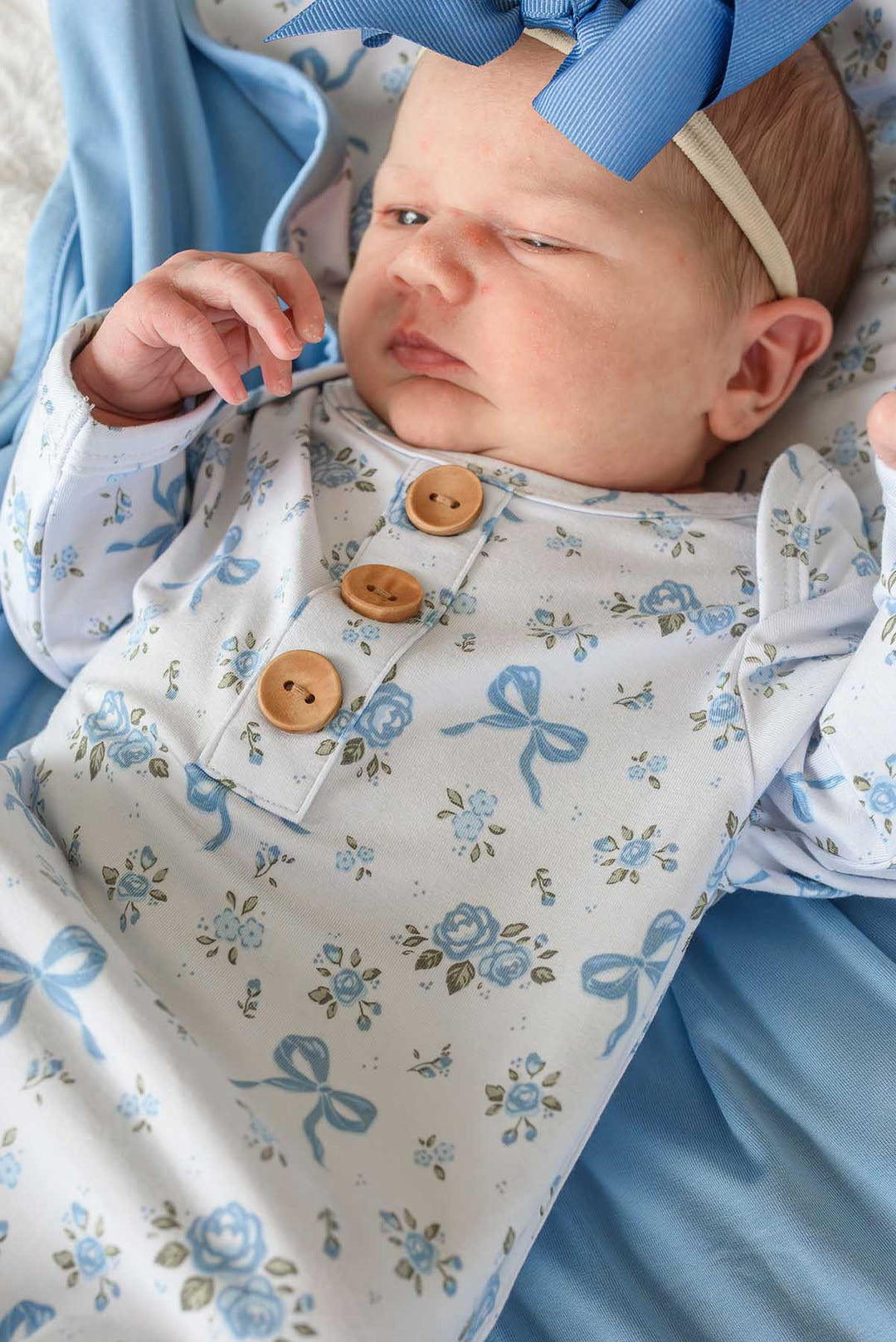 Baby with a blue bow headband wearing a Tied In Blooms knot gown with blue floral and bow print and wooden buttons