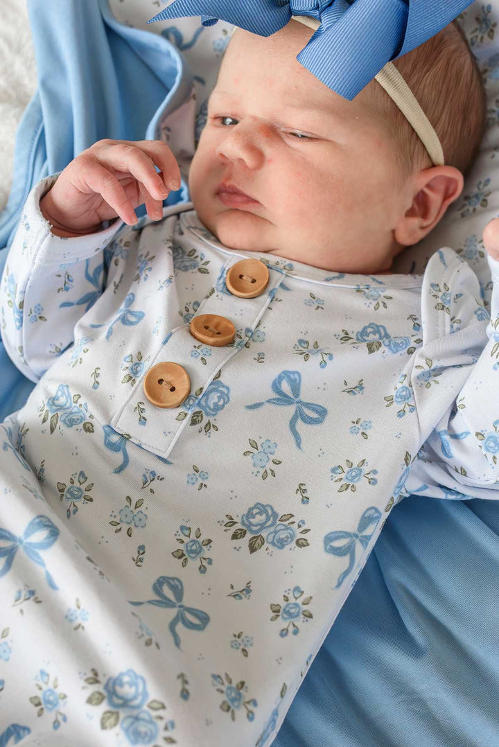 Baby with a blue bow headband wearing a Tied In Blooms knot gown with blue floral and bow print and wooden buttons