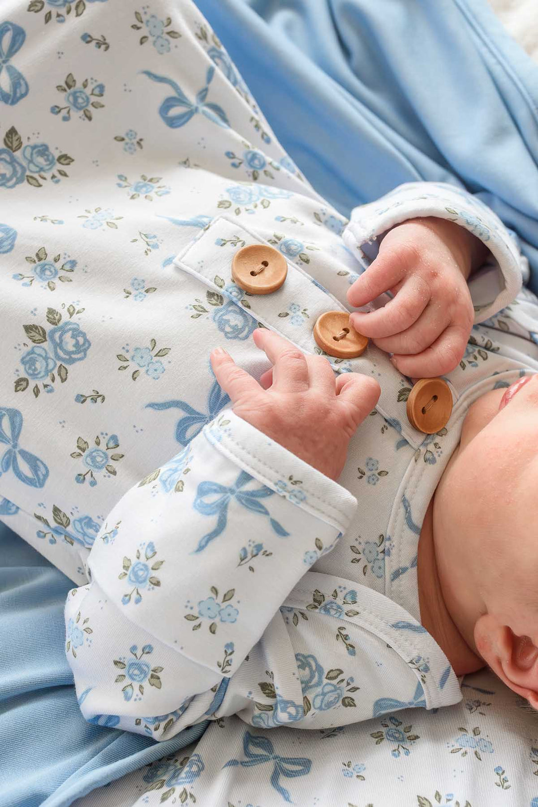 Close-up of a baby's hands on the wooden buttons of a Tied In Blooms knot gown in blue floral and bow print