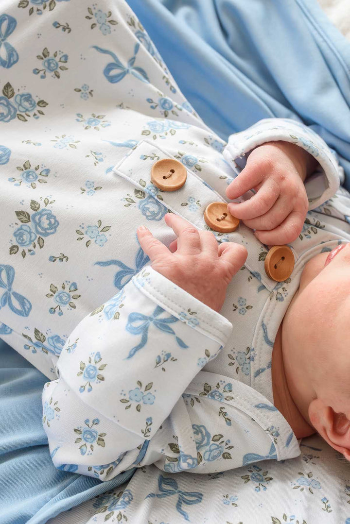 Close-up of a baby's hands on the wooden buttons of a Tied In Blooms knot gown in blue floral and bow print