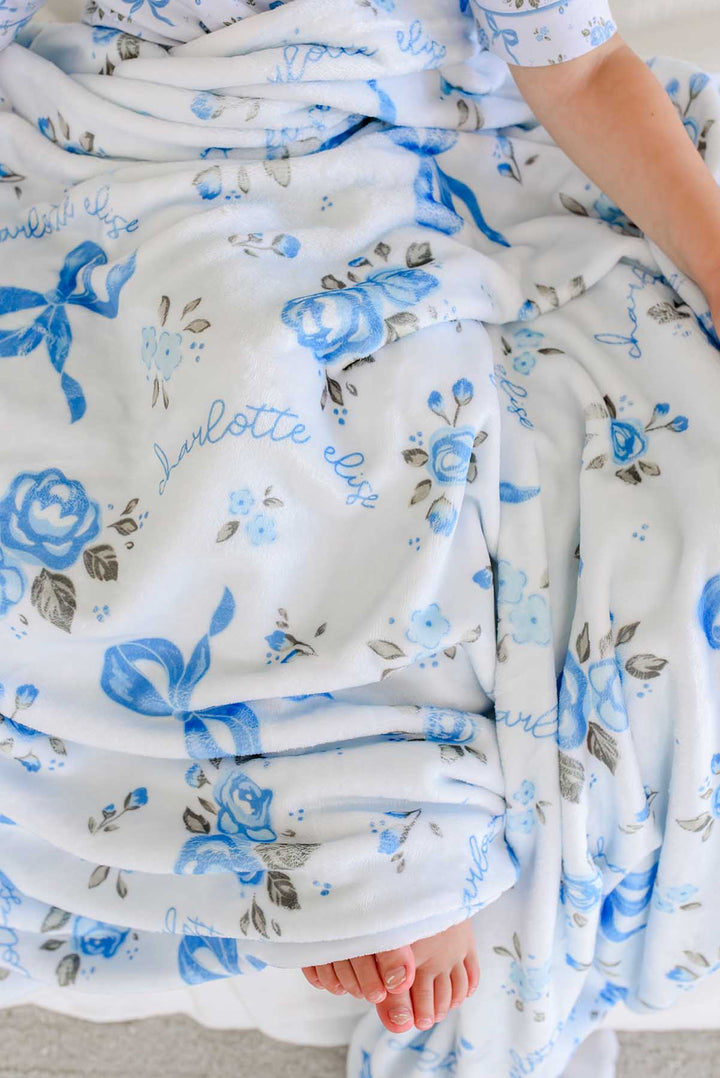 Close-up of a child wrapped in a Tied In Blooms personalized blanket with blue floral and bow print, showing the name Charlotte Elise in cursive script