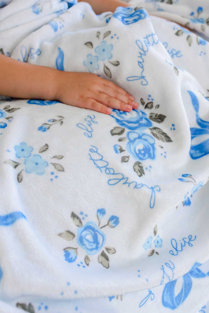 Close-up of a child's hand resting on a Tied In Blooms personalized blanket with blue floral and bow print and a name in cursive script
