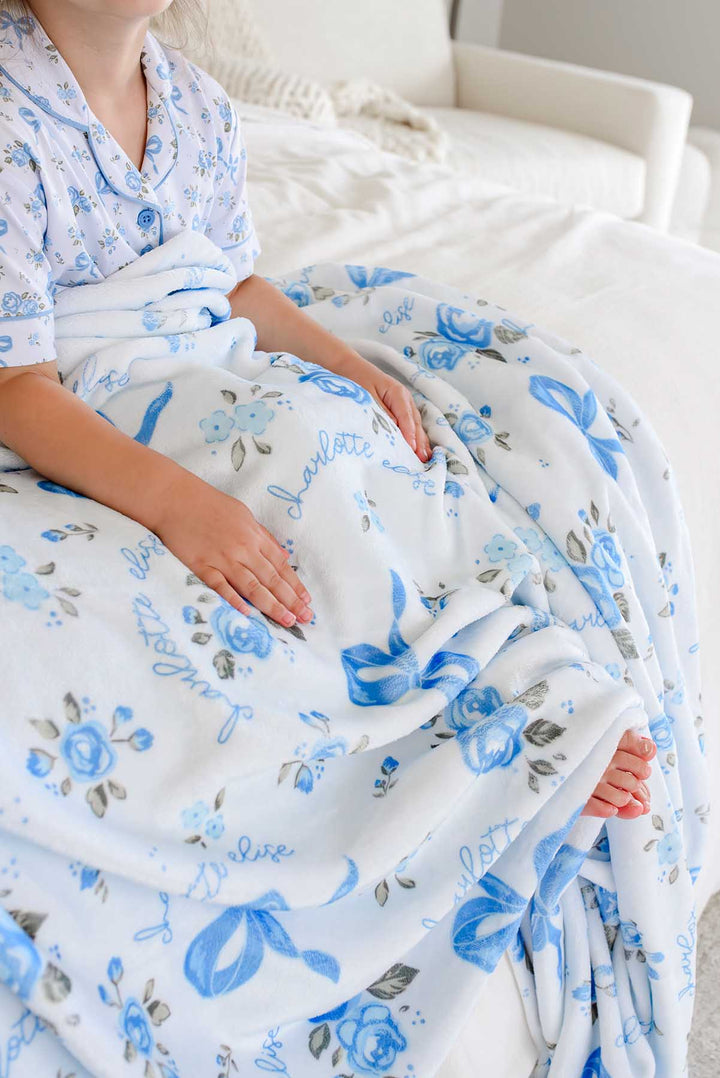Child sitting on a bed with hands resting on a Tied In Blooms personalized blanket in blue floral and bow print, a bare foot peeking out