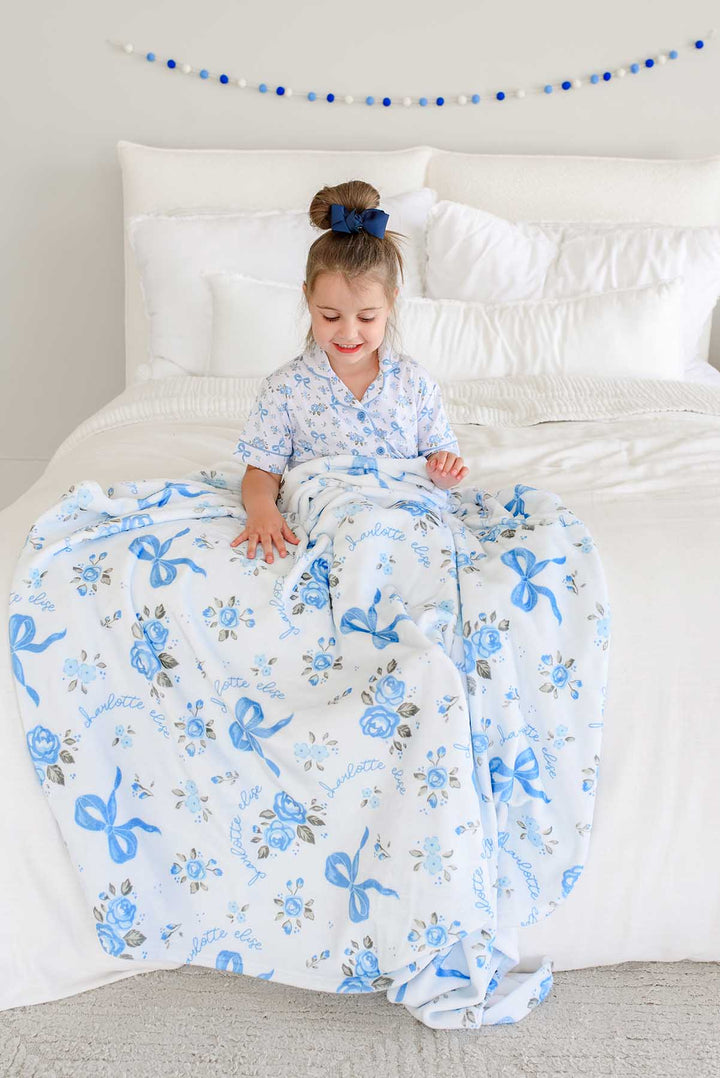 Little girl sitting on a bed in a nursery, spreading out a Tied In Blooms personalized blanket in blue floral and bow print