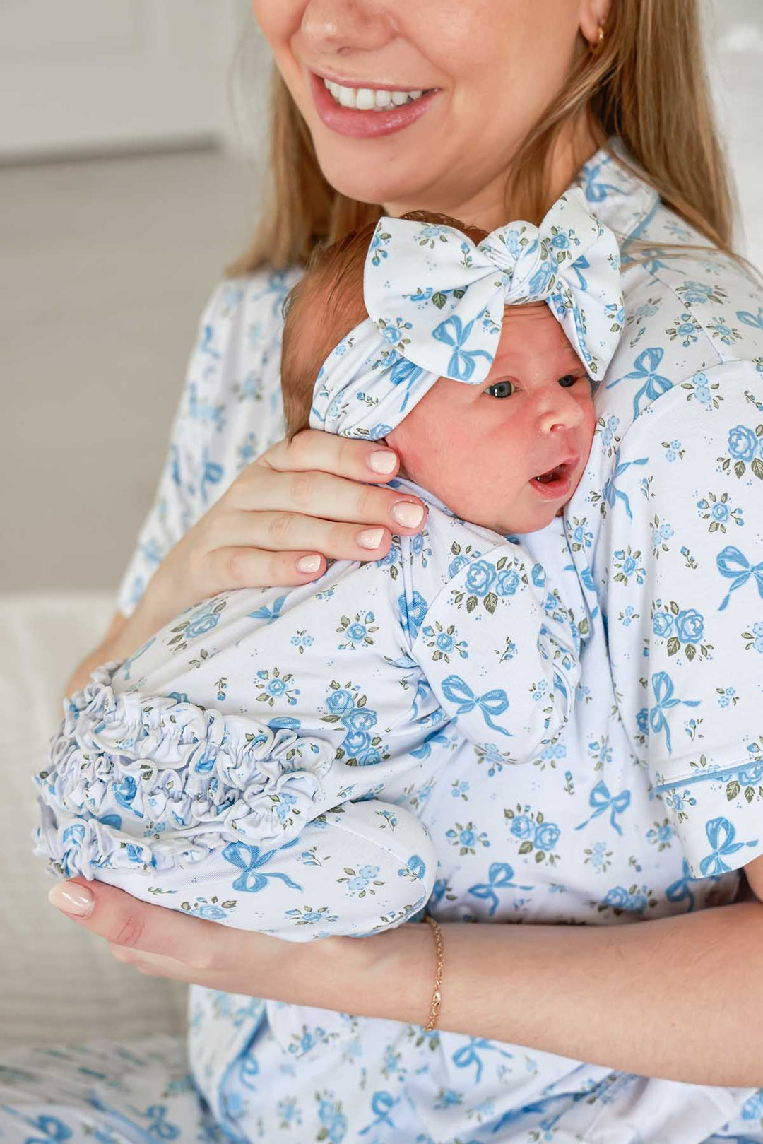 Smiling mother holding her baby, who wears a Tied In Blooms bow headwrap and ruffled outfit in blue floral and bow print