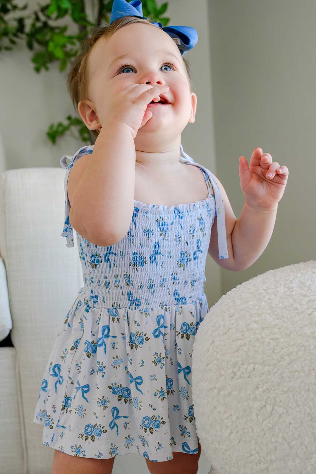 Curious toddler with a blue bow headband wearing a Tied In Blooms smocked twirl bodysuit in blue floral and bow print