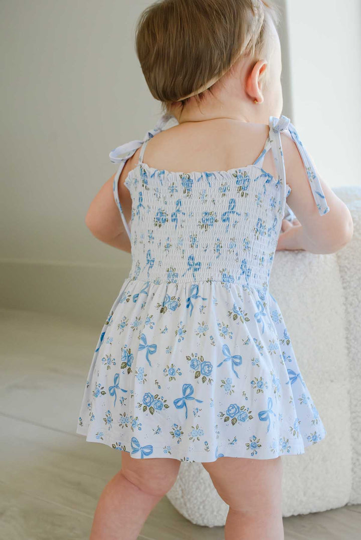 Back view of a toddler wearing a Tied In Blooms smocked twirl bodysuit in blue floral and bow print, showing the tie straps and twirl skirt