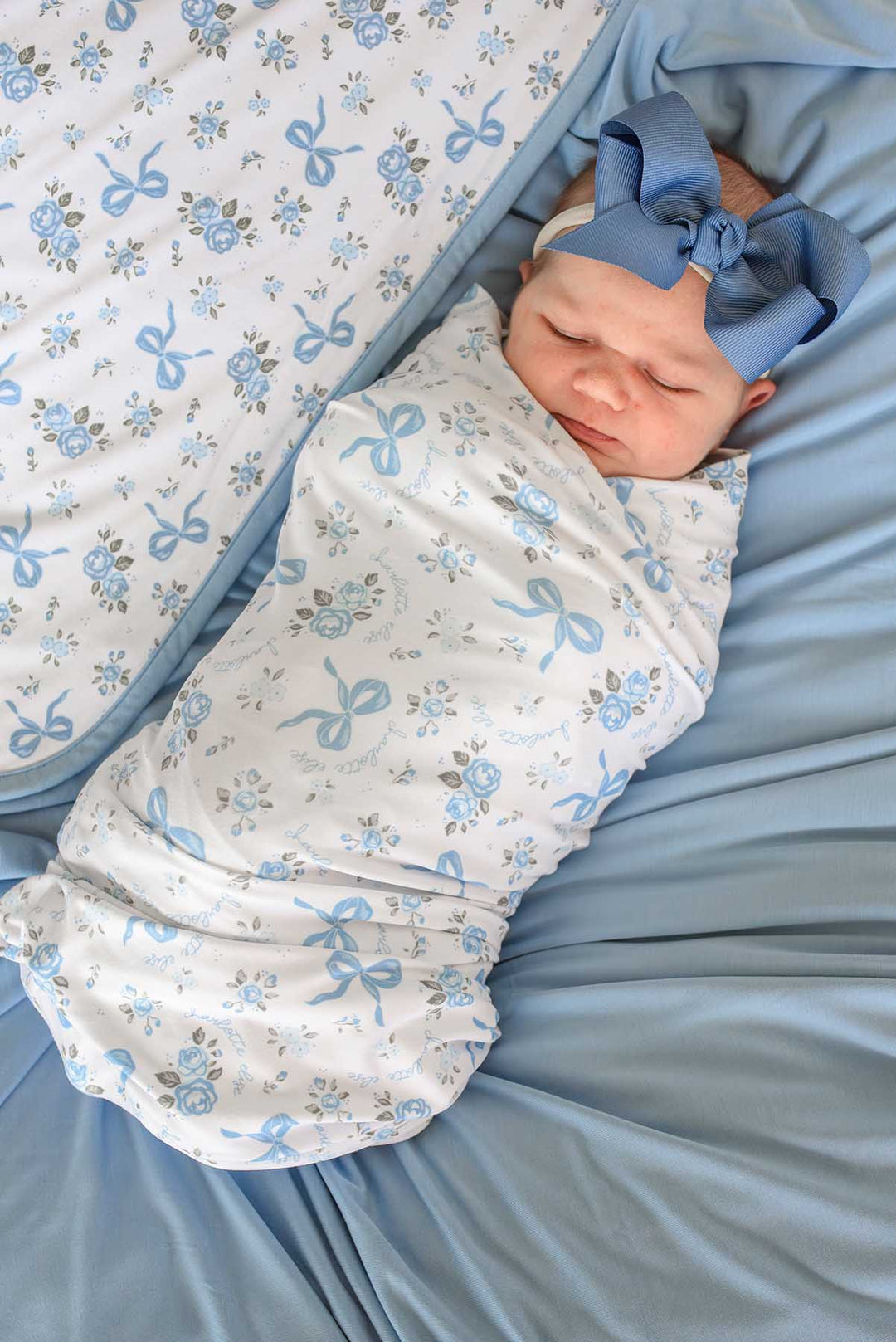 Sleeping newborn baby wearing a blue bow headband, swaddled in a Tied In Blooms blanket with blue floral and bow print