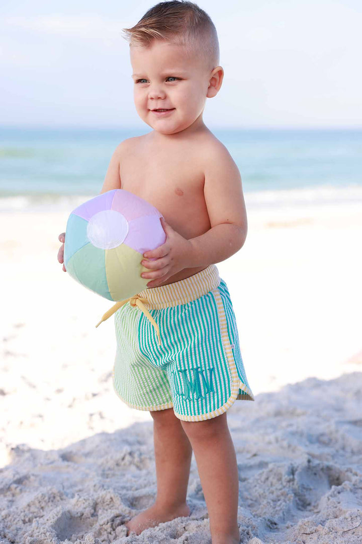 boy standing on the beach in multicolor seersucker swim trunks holding a beach ball 