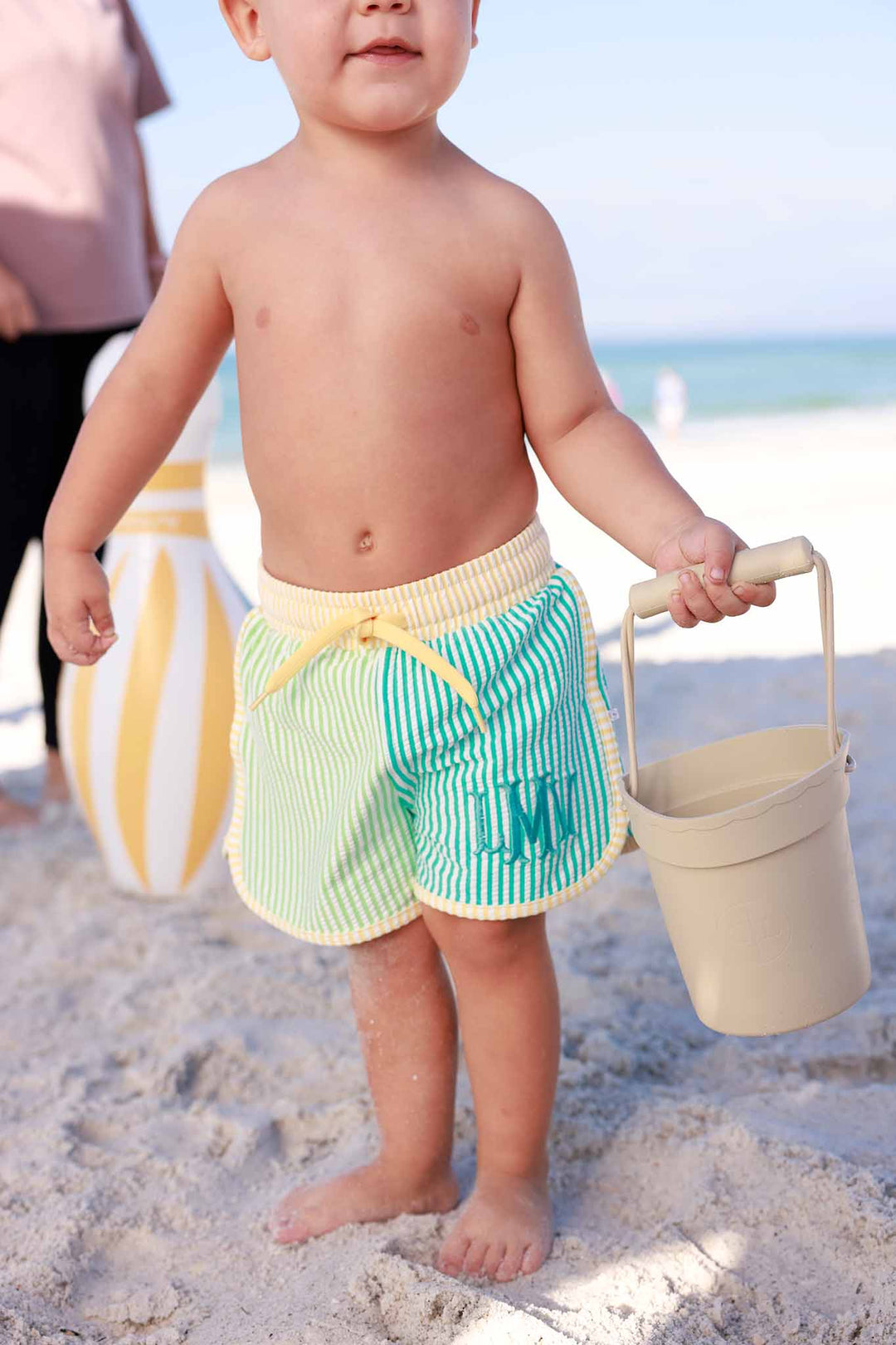 baby boy wearing color block swim trunks holding a neutral beach bucket 