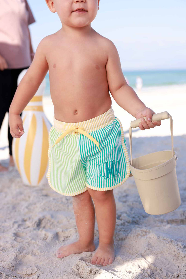 baby boy wearing color block swim trunks holding a neutral beach bucket 