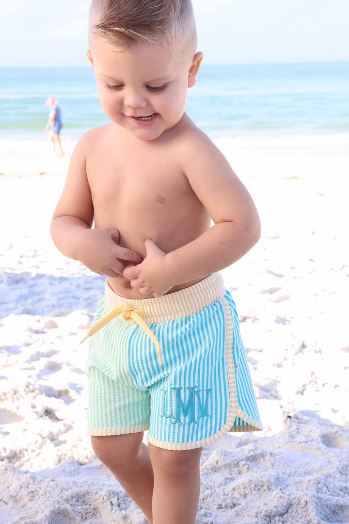 toddler boy playing on the beach in blue, green and yellow swim trunks 
