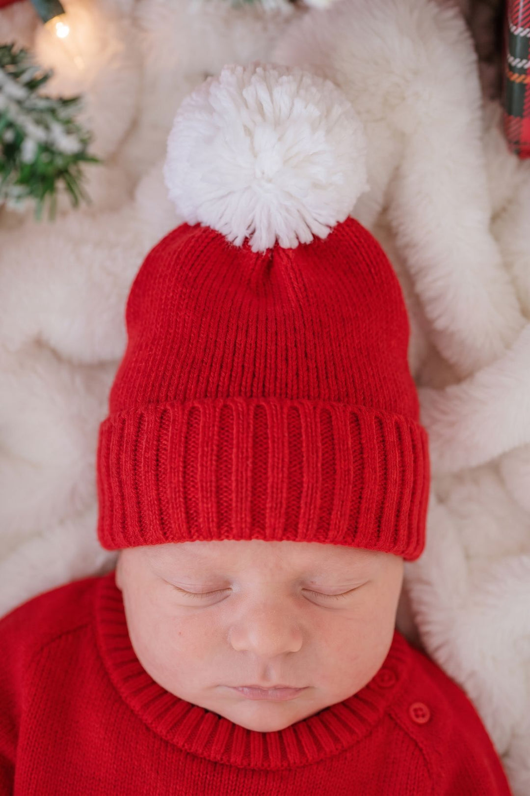 Close-up of a sleeping newborn wearing a Caden Lane red knit beanie with a white pom-pom, paired with a red knit sweater.