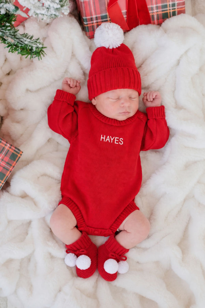 Sleeping newborn wearing a Caden Lane red knit beanie with a white pom-pom and a personalized red knit bodysuit reading "Hayes," paired with red knit booties with white pom-poms.