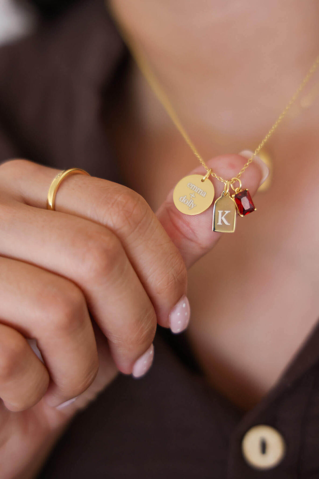 Caden Lane gold necklace held in hand featuring engraved "emma + dolly" disc, initial "K" tag charm, and red birthstone charm