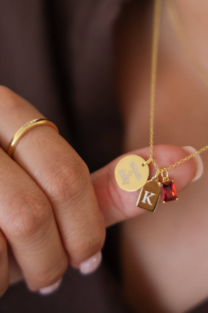 Caden Lane gold necklace held in hand featuring engraved "emma + dolly" disc, initial "K" tag charm, and red birthstone charm, with matching stackable ring