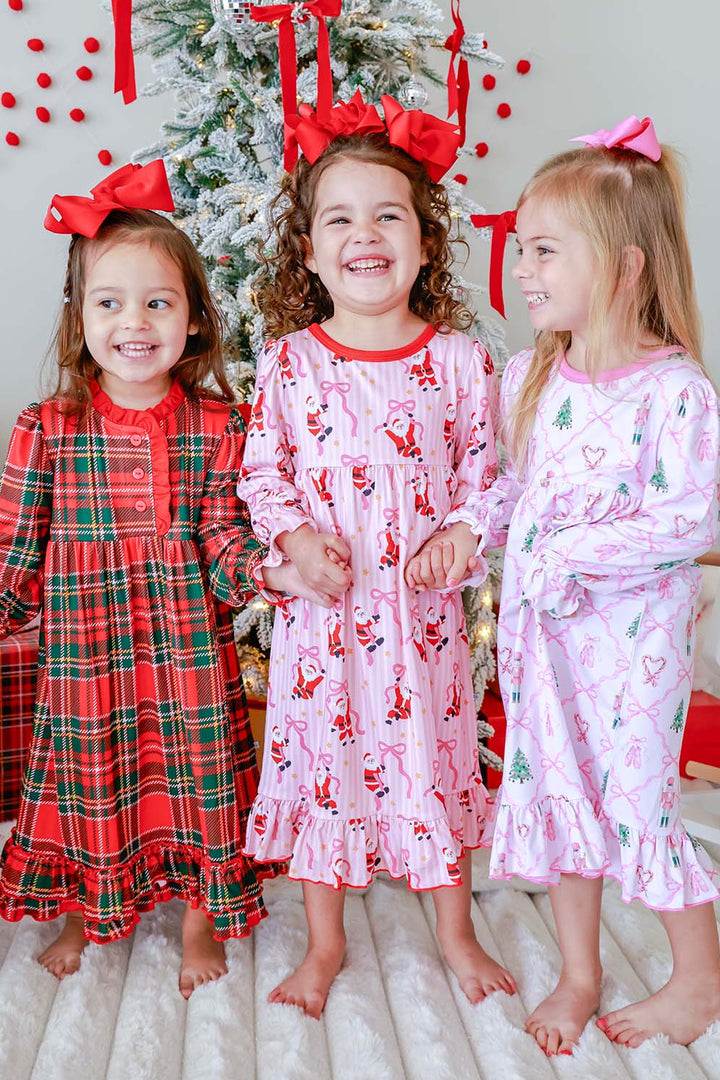 Three girls in matching Caden Lane Christmas nightgowns — red plaid, pink Santa print, and pink nutcracker print — standing by a flocked Christmas tree