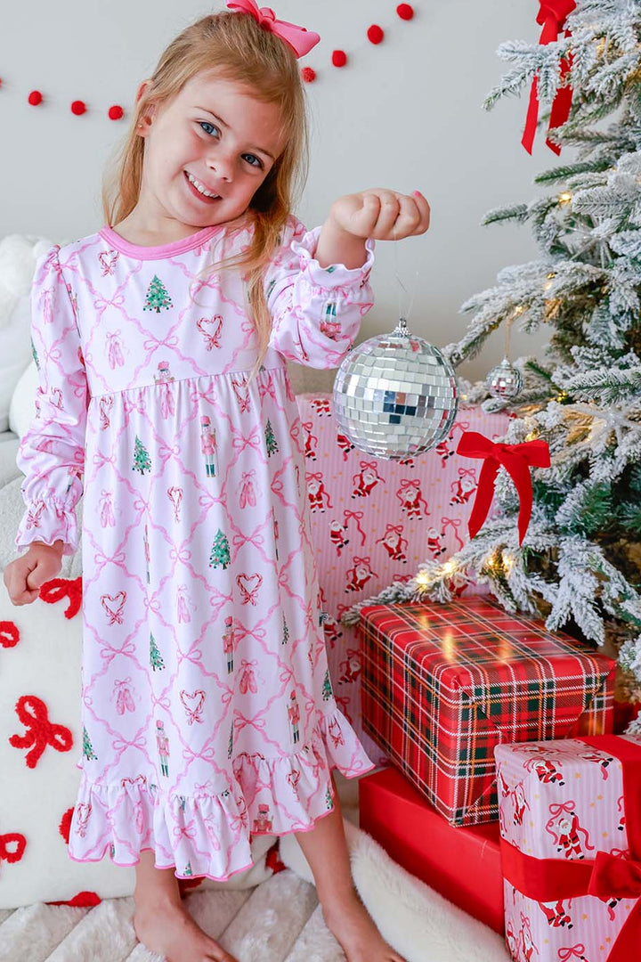 Girl in Caden Lane pink nutcracker-print nightgown holding a disco ball ornament by the Christmas tree
