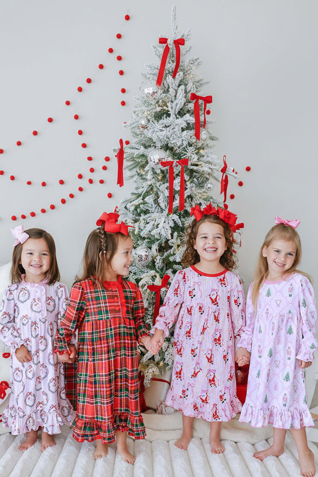 Four girls holding hands in matching Caden Lane holiday nightgowns — wreath print, red plaid, Santa print, and nutcracker print — in front of a decorated tree