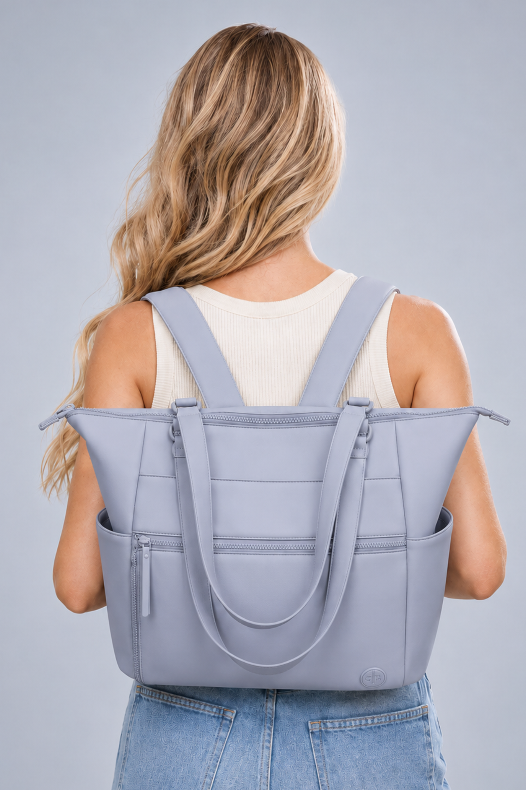 A woman viewed from behind wearing a light blue-gray tote bag over her shoulders. The bag has dual shoulder straps, a zip-top closure, and multiple front pockets.