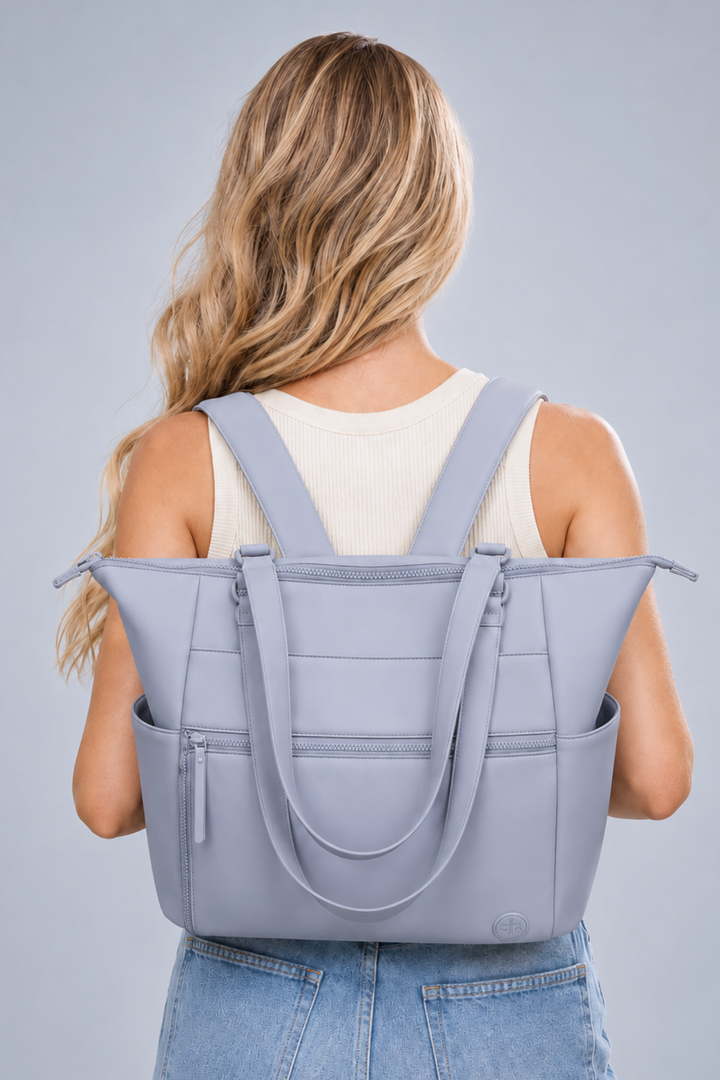 A woman viewed from behind wearing a light blue-gray tote bag over her shoulders. The bag has dual shoulder straps, a zip-top closure, and multiple front pockets.