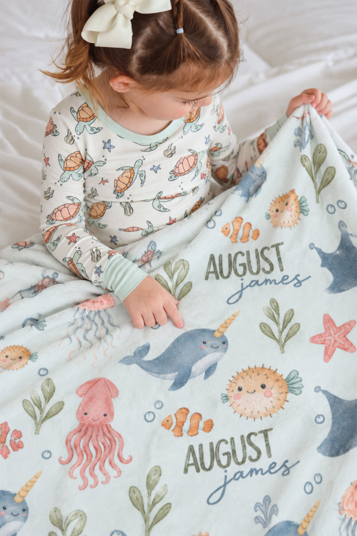 Girl in sea turtle-print pajamas sitting on a bed touching a personalized Caden Lane Ocean Dreams blanket, showing narwhal, pufferfish, and custom name print