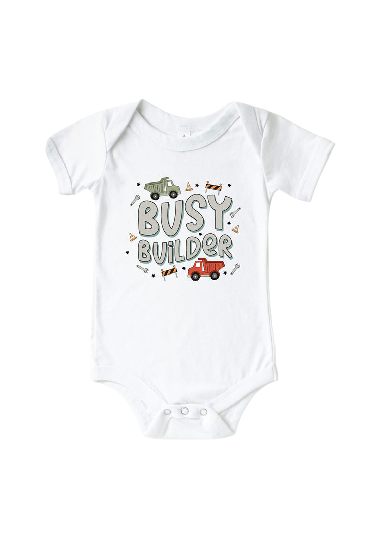 Flat lay of a Caden Lane white short-sleeve bodysuit in a Busy Builder print featuring a green and a red dump truck, cones, and wrenches, reading "Busy builder."
