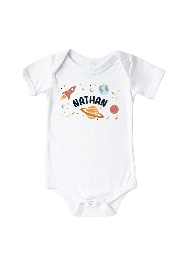Flat lay of a Caden Lane white short-sleeve bodysuit with a Stellar Snuggles Planets graphic featuring labeled planets, two rockets, and scattered stars.