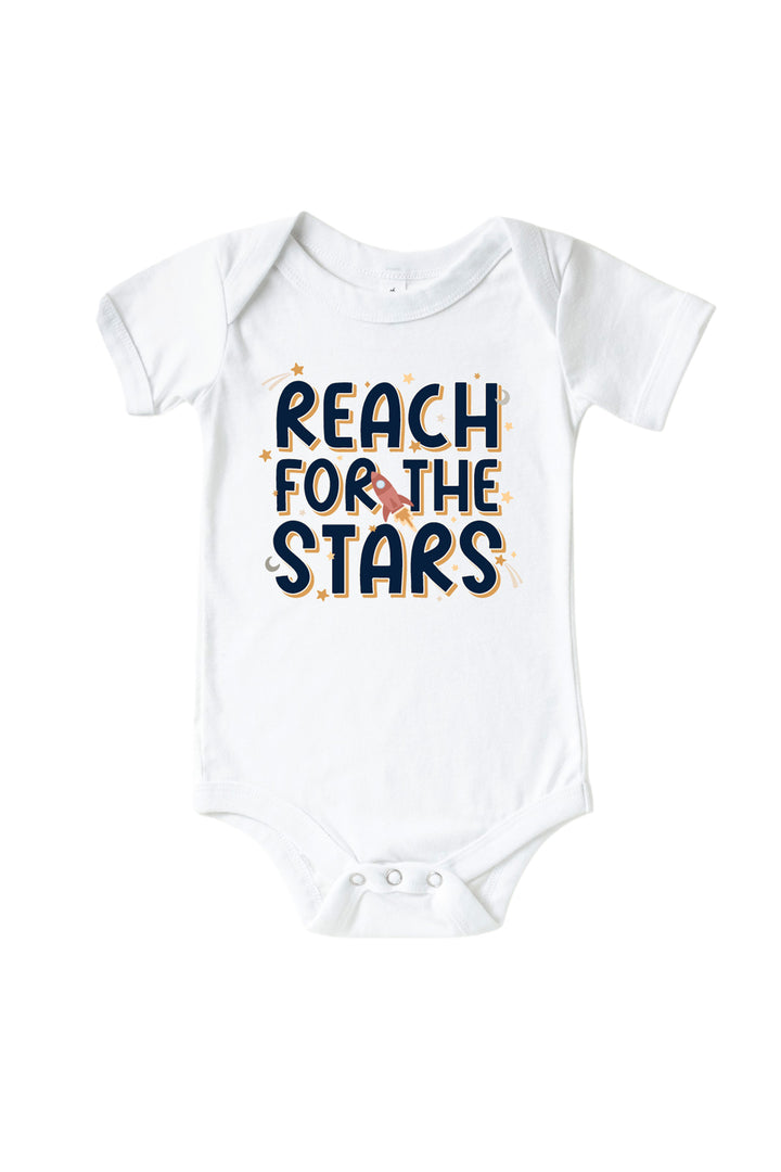 Flat lay of a Caden Lane white short-sleeve bodysuit with a Stellar Snuggles graphic featuring a rocket and stars, reading “Reach for the Stars.”