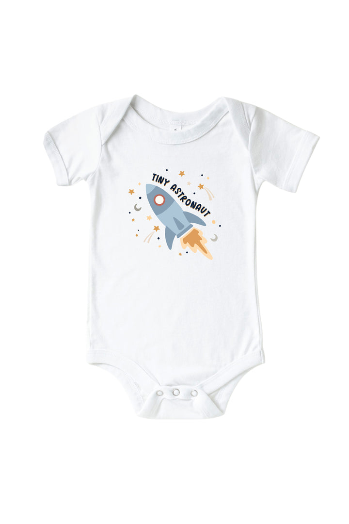 Flat lay of a Caden Lane white short-sleeve bodysuit with a Stellar Snuggles graphic featuring a blue rocket among stars and moons, reading “Tiny Astronaut.”