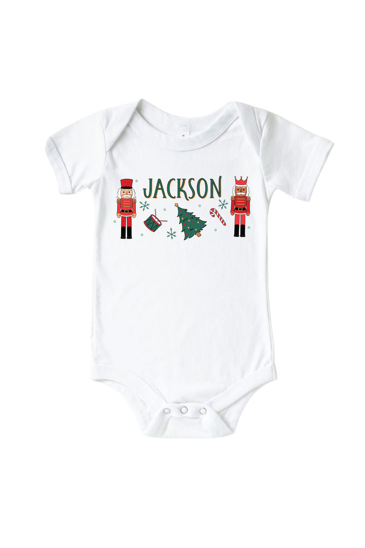 Caden Lane's white toddler t-shirt personalized with the name "Jackson," featuring red toy soldier nutcrackers, a Christmas tree, drum, and candy cane, laid flat.