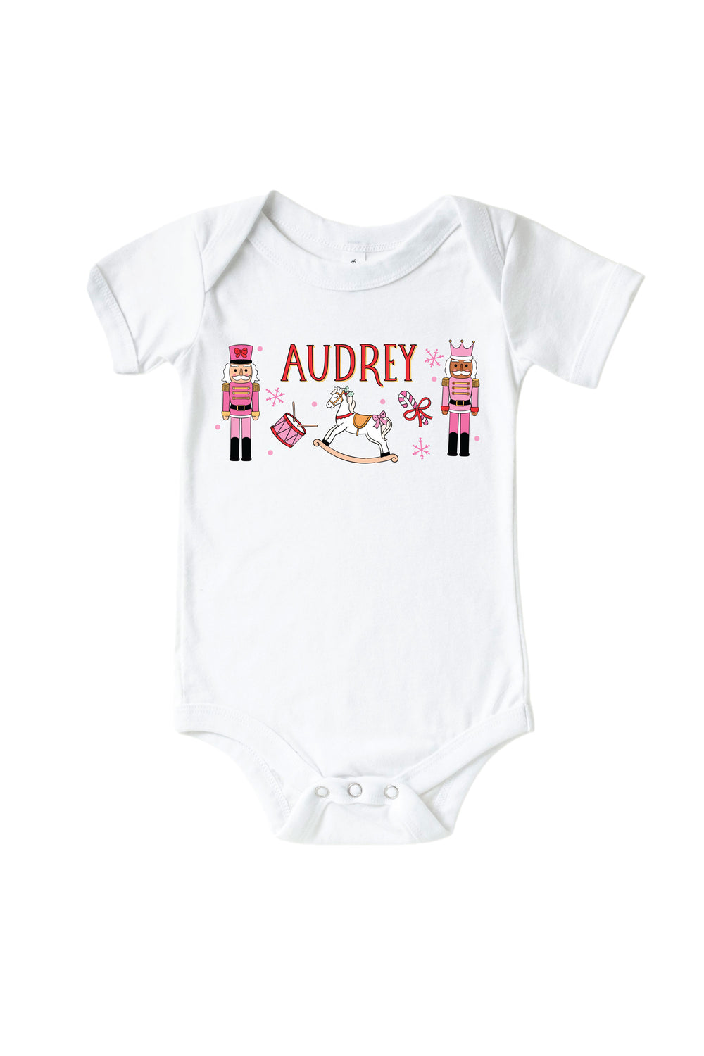 Caden Lane's white baby onesie personalized with the name "Audrey," featuring pink toy soldier nutcrackers, a rocking horse, drum, and candy cane, laid flat.