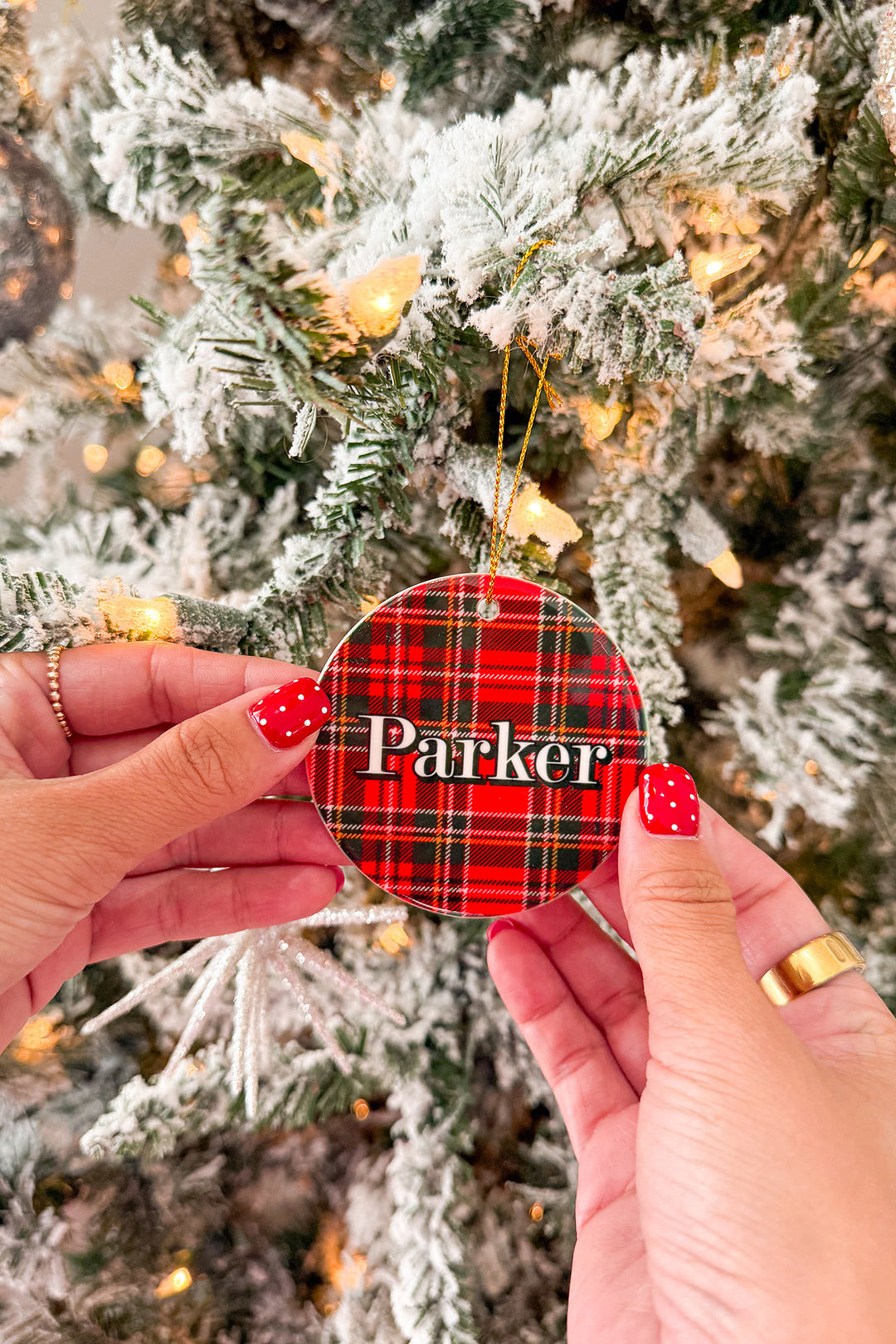 A round Caden Lane Christmas ornament with a red, green, and black plaid pattern and the name 'Parker' in white, held by two hands in front of a decorated flocked Christmas tree.